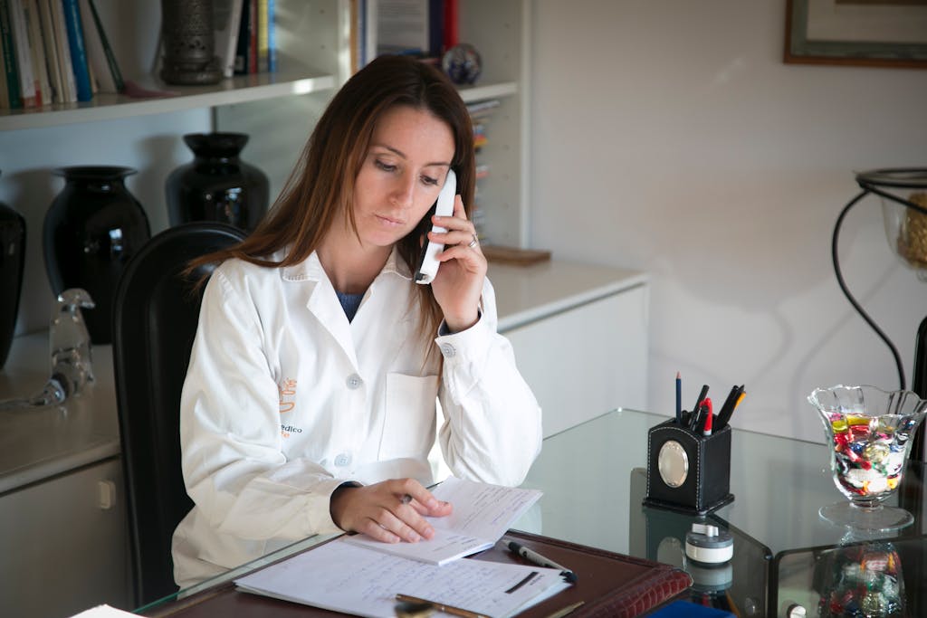 A female doctor engaged in a phone call while taking notes at her office desk.